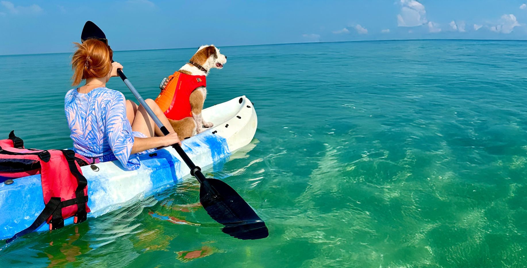 Woman paddling a kayak with her brown and white dog in a red life jacket, looking out over the turquoise ocean during a pet-led journey.