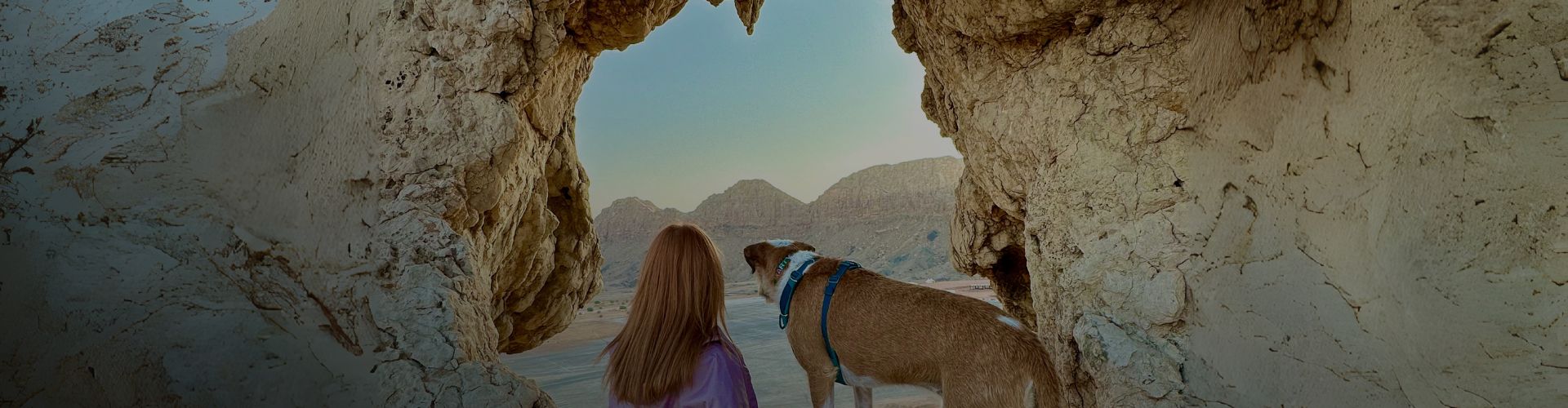 Woman and her dog framed by a rocky cave opening, overlooking desert mountains at sunrise — capturing the spirit of pet-led adventures.