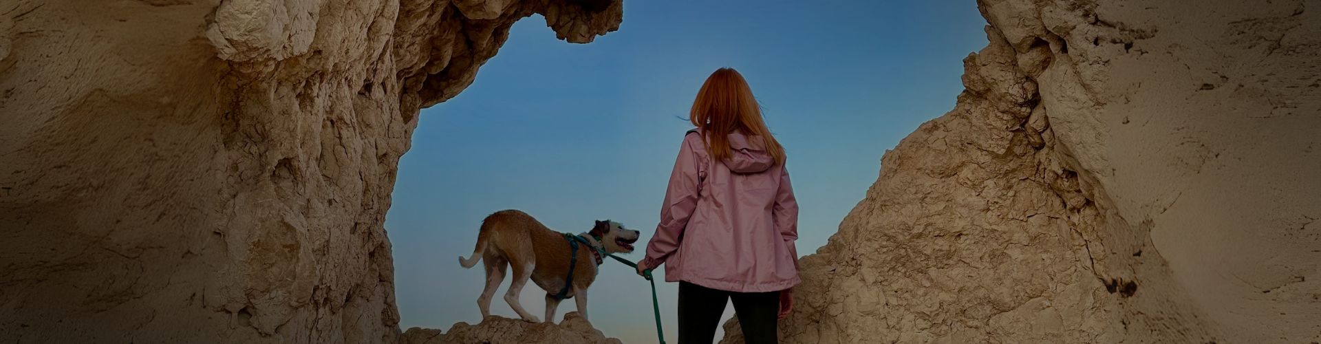 Woman in a pink jacket standing with her rescue dog on a rocky cliff, looking out through a natural stone arch at the mountains ahead — symbolizing adventure and pet-led travel.
