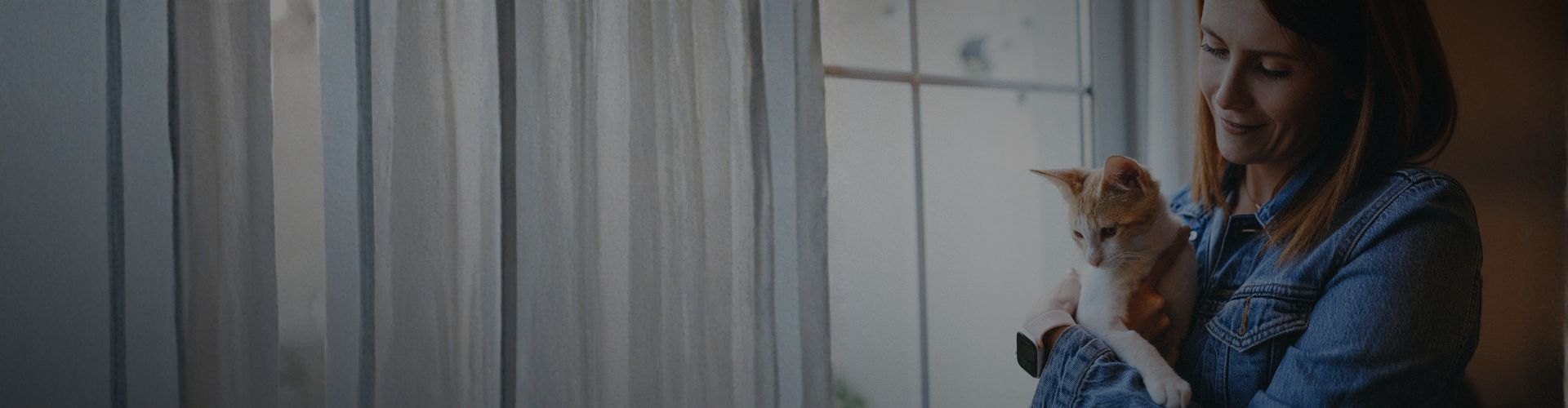Carmen holding an orange-and-white cat near a softly lit window with curtains.